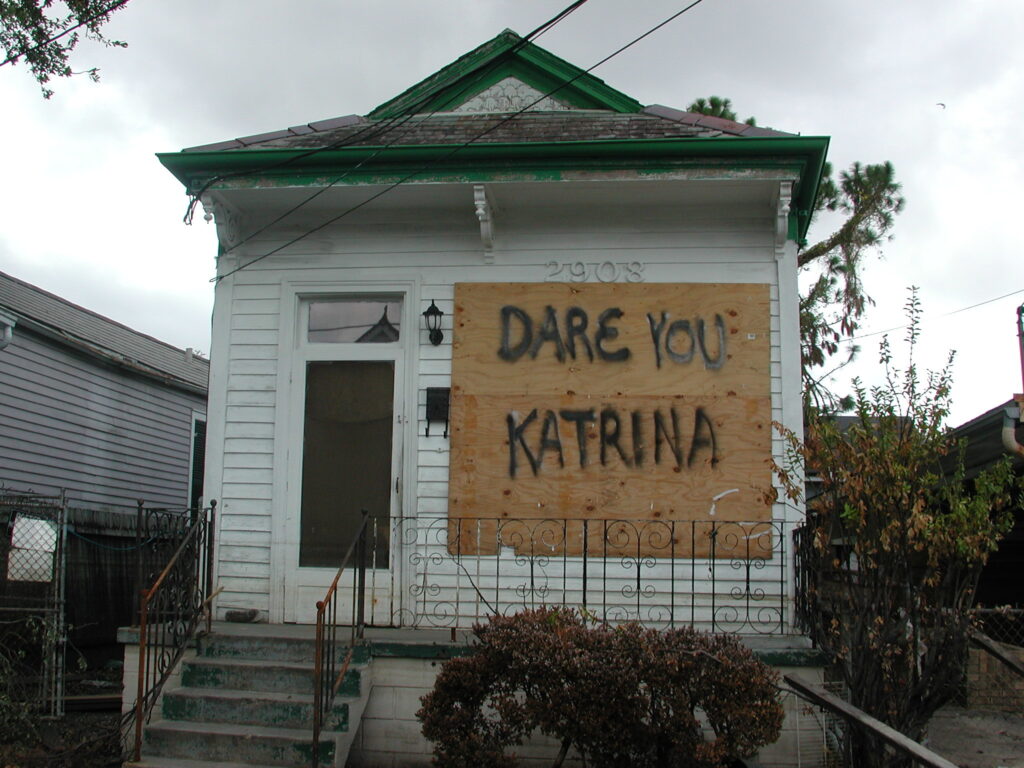 Photo of a plywood board affixed to the front of a house with the words "Dare You Katrina" spray-painted